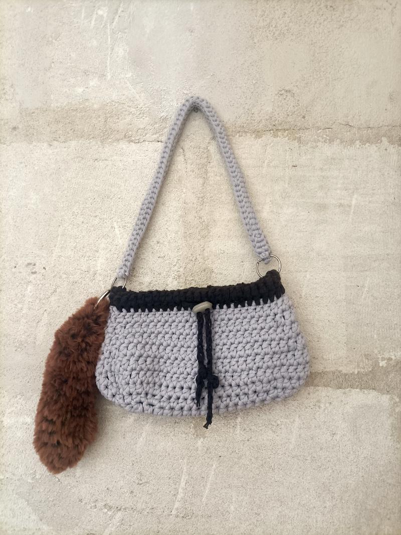 sac en crochet gris et marron avec un porte clés en fausse fourrure rousse posé sur un banc en bois sur un fond naturel avec des feuilles mortes et arbres, un livre dépasse du sac