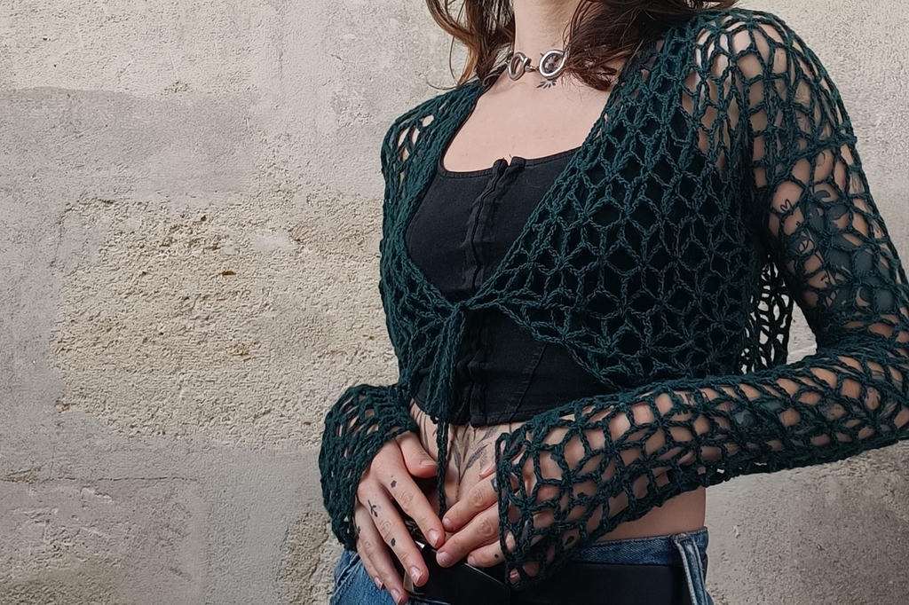 jeune femme tatouée portant un boléro en crochet vert émeraude au point ajouré et aux motifs de fleurs sur fond pierre