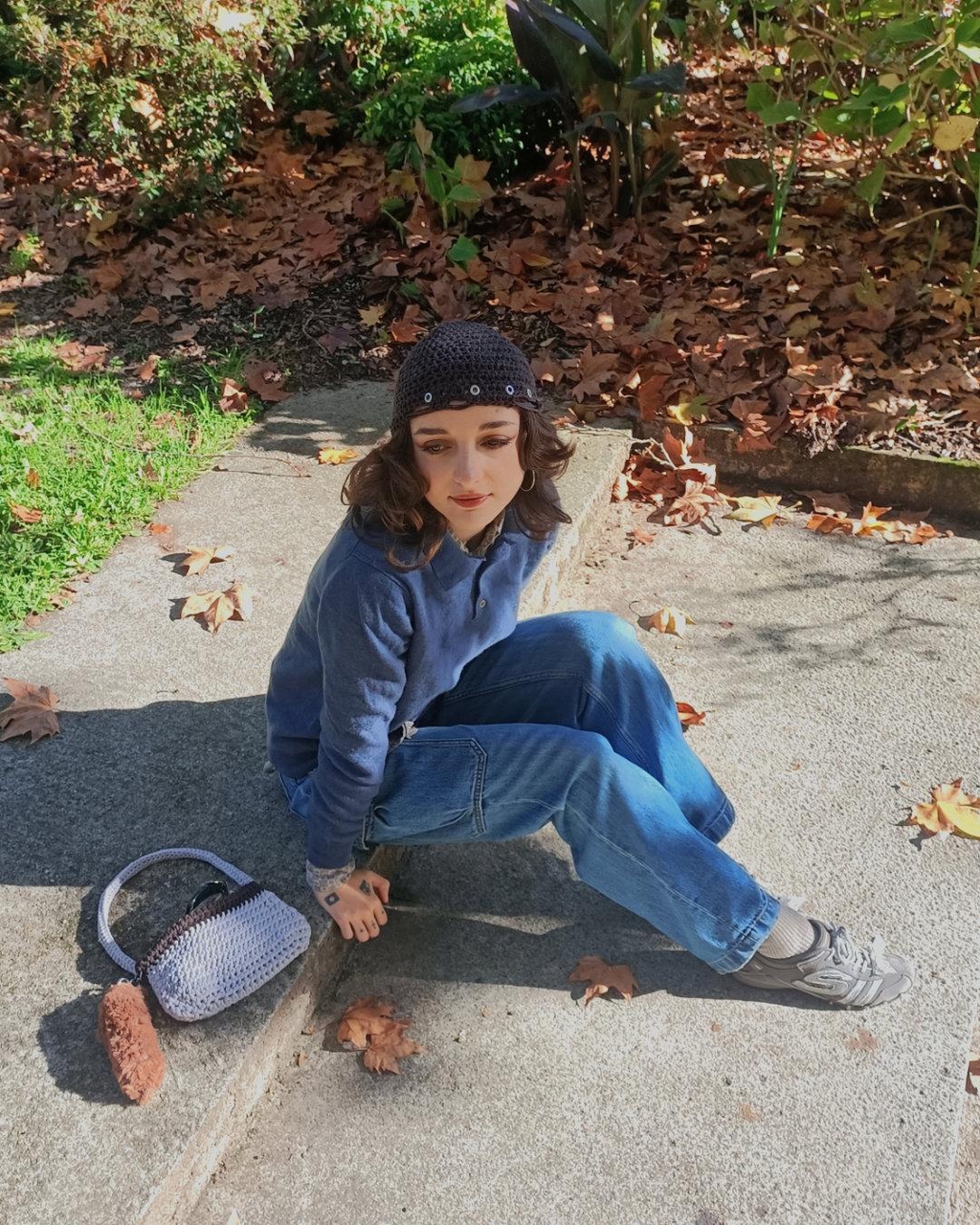 femme tatouée assise par terre sur un sol en béton, portant un jean, un pull et un bonnet en crochet marron avec des œillets, un sac en crochet gris et marron avec un porte clés en fausse fourrure est posé à côté d'elle. Décor automnal avec des feuilles mortes et lumière chaleureuse
