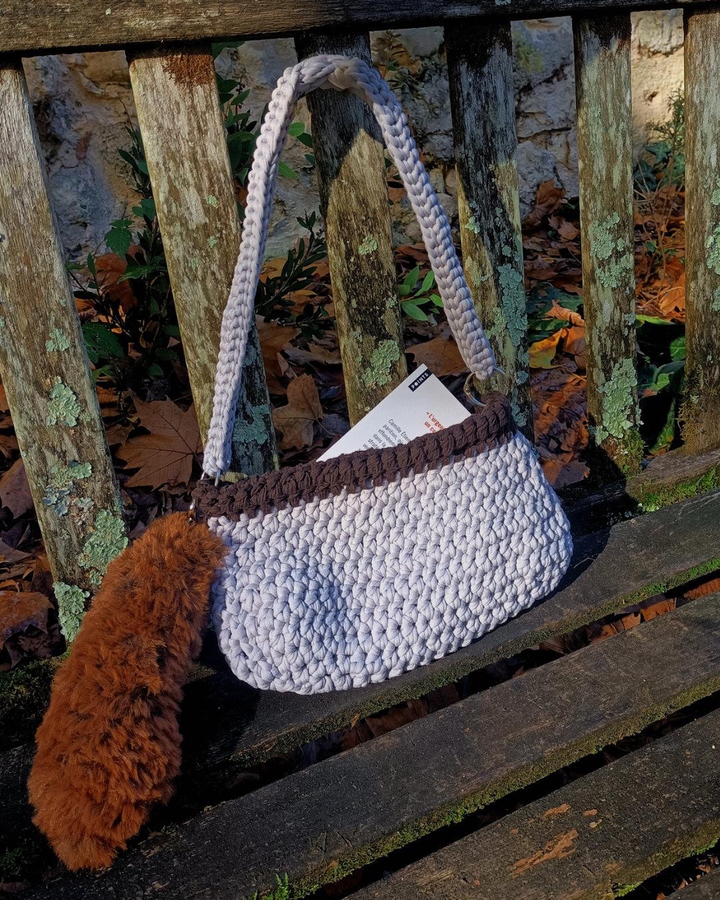 sac en crochet gris et marron avec un porte clés en fausse fourrure rousse posé sur un banc en bois sur un fond naturel avec des feuilles mortes et arbres, un livre dépasse du sac