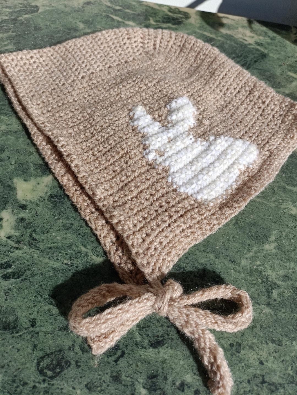 bonnet en crochet beige avec un motif de lapin blanc sur le côté, posé sur une table en marbre verte