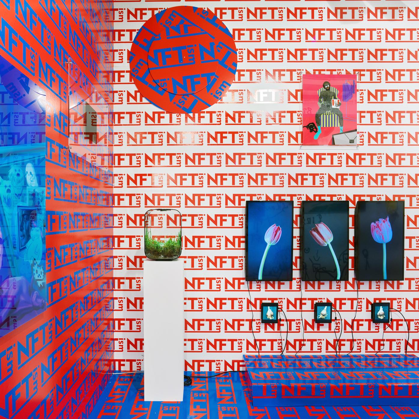 An Exhibition view with digital artworks by Sarah Friend, Anna Ridler, Olive Allen, Osinachi and a wallpaper by Kenny Schachter reading "NFTism" multiple times in red, white and blue.