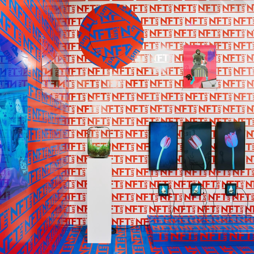 An Exhibition view with digital artworks by Sarah Friend, Anna Ridler, Olive Allen, Osinachi and a wallpaper by Kenny Schachter reading "NFTism" multiple times in red, white and blue. 