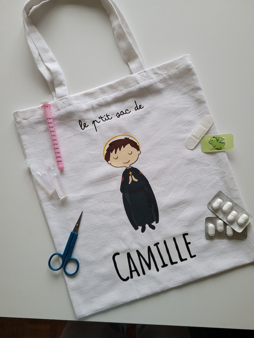 Photo d'un totebag avec la phrase " le p'tit sac de Camille" et un dessin de Saint Camille de Lellis. Des médicaments, pipettes et pansements en guide de décoration autour du sac car saint Camille est le saint patron des infirmiers, malades..