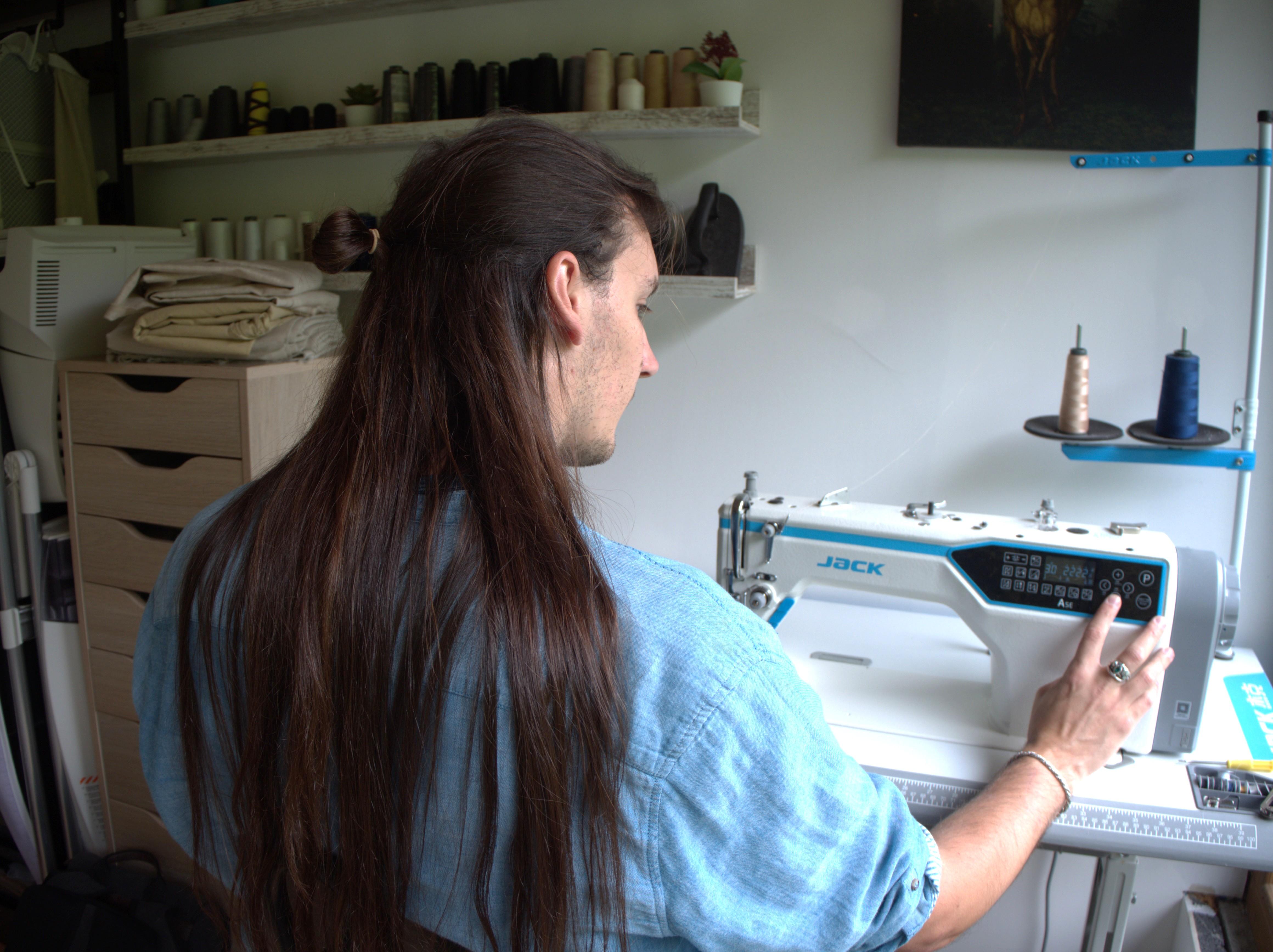 Photo de L'Atelier des Enchanteurs, montrant Alexandre PETROD assis devant la piqueuse plate, les stocks de tissus et de bobines de fil de couture, ainsi que deux fers à repasser anciens. 