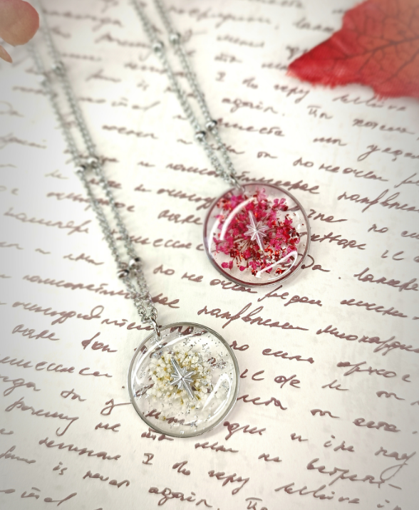 2 colliers au médaillon rond sont posés sur une feuille manuscrite dans un esprit vintage. Ils sont en acier, renferment de vraies fleurs séchées, des paillettes et une étoile du Nord au Centre pour symboliser le guide.