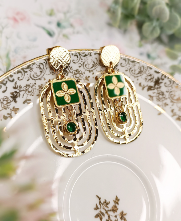 Une paire de boucles d'oreilles pendantes, en acier doré et cristal vert. Breloque ovale avec un motif géométrique martelé ajouré. 