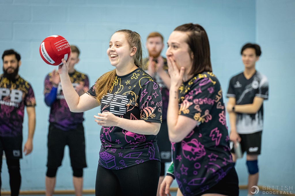 PB Dodgeball competitive team, the photo is focussed on two women at the foreground, one is smiling and throwing a dodgeball, the other has their hand over their mouth, indicating something shocking has happened, outside of the frame.