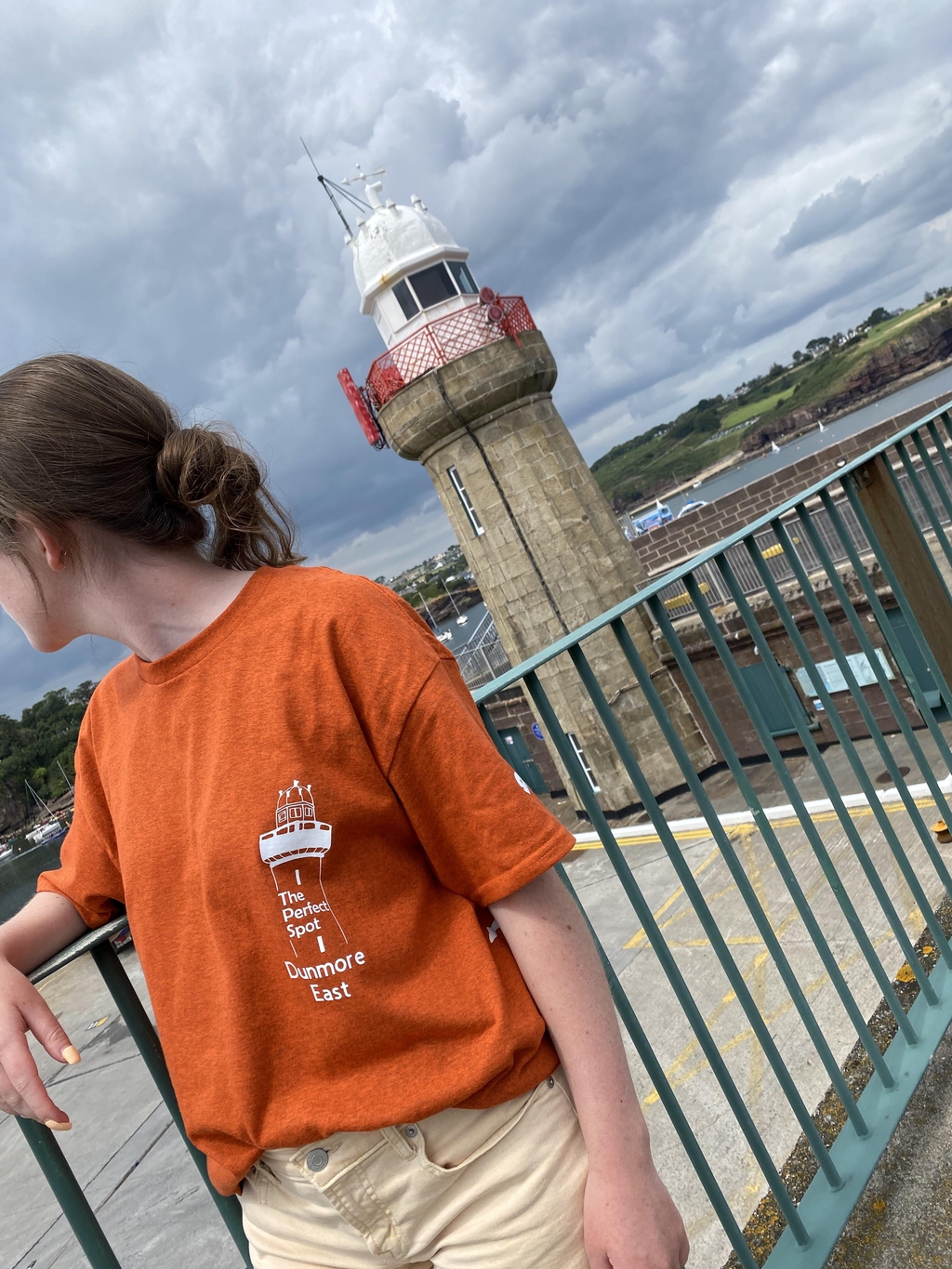pictured in a woman with soft brown hair tied in a bun she is turned to look away from the camera wearing a bright orange t-shirt we can see it features a white graphic of the lighthouse with The perf et spot inscribed inside the graphic written below it is dunmore east the same lighthouse is pictured behind her. 