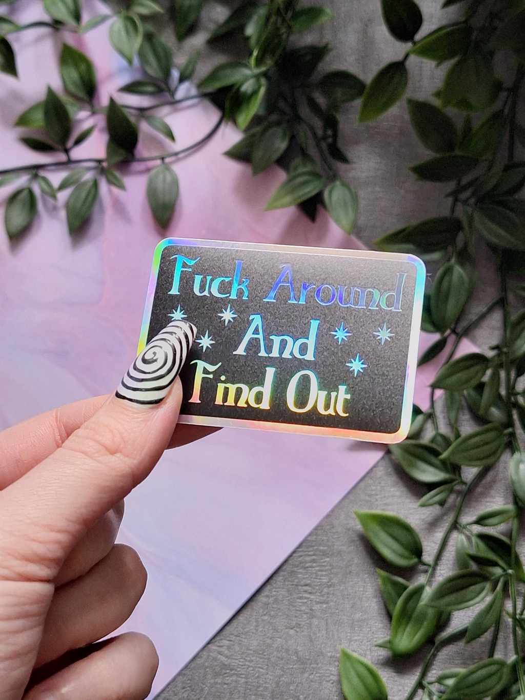 The woman is holding a holographic and matt black sticker showing the words Fuck Around and find out. This selection is for all the stickers including large, small, holographic and pride representation.