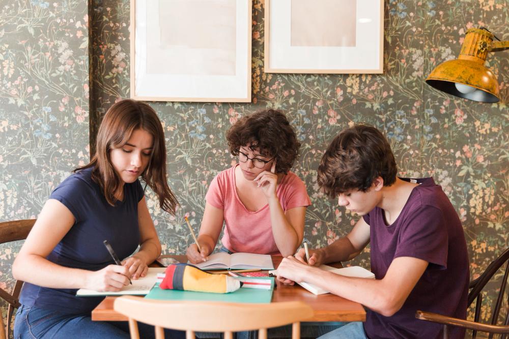 Groupe de trois jeunes concentrés en train d’écrire autour d’une table en bois, avec un décor fleuri en arrière‑plan.