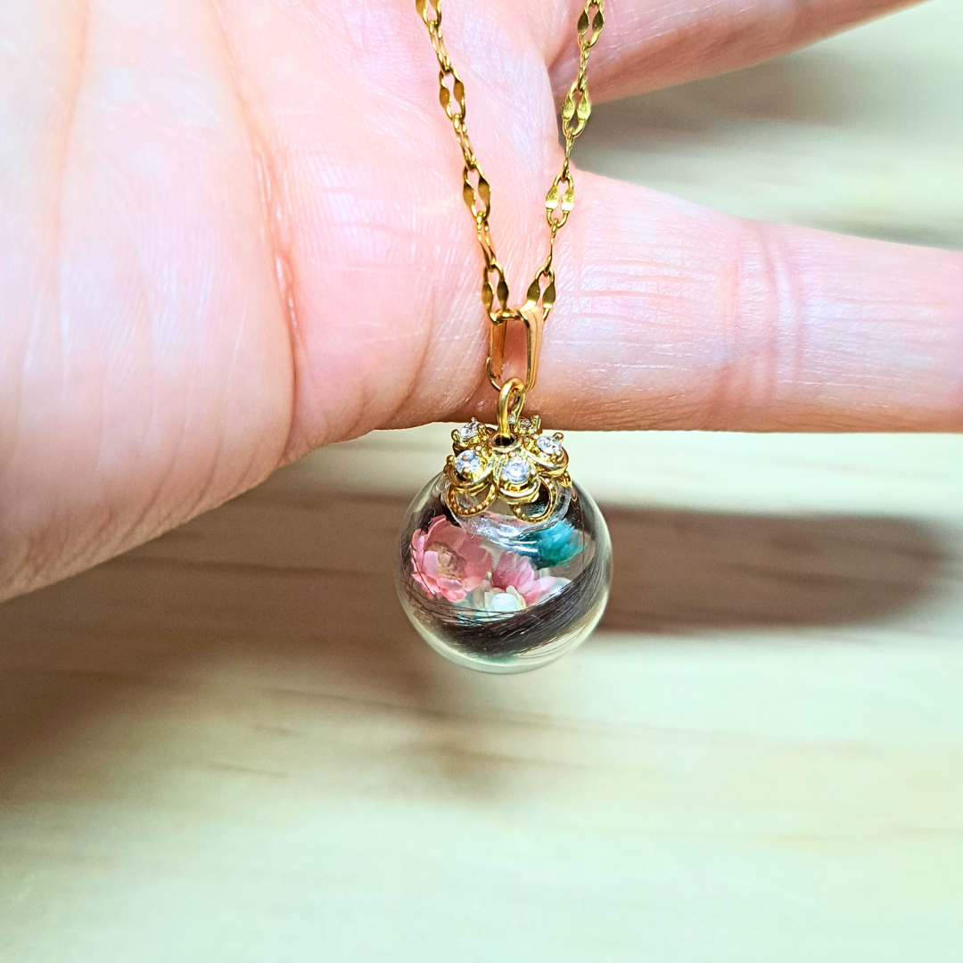 Bijou hommage de créateur : collier chaîne acier inoxydable plaqué doré, pendentif boule de verre soufflé transparent incluant mèche de cheveux, fleurs séchées blanches, rose pastel et vert pastel & perles de nacre blanche