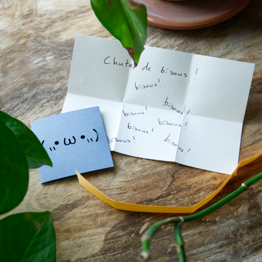 Une mini-carte en pochon présentant un kaomoji découpé au laser. Un petit papier est glissé dedans et se retire à l'aide d'un ruban, prêt à recevoir votre écriture pour y cacher un petit mot doux.