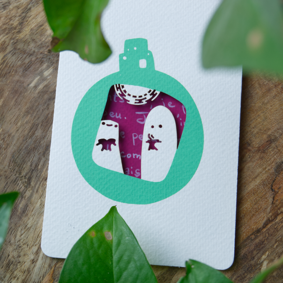 Une carte sérigraphiée présente deux personnages au centre d'une boule de Noël vert sauge : l'un tient un livre, l'autre une tasse de café, et un luminaire les éclaire par-dessus. La carte est ajourée et laisse voir l'intérieur couleur cassis.