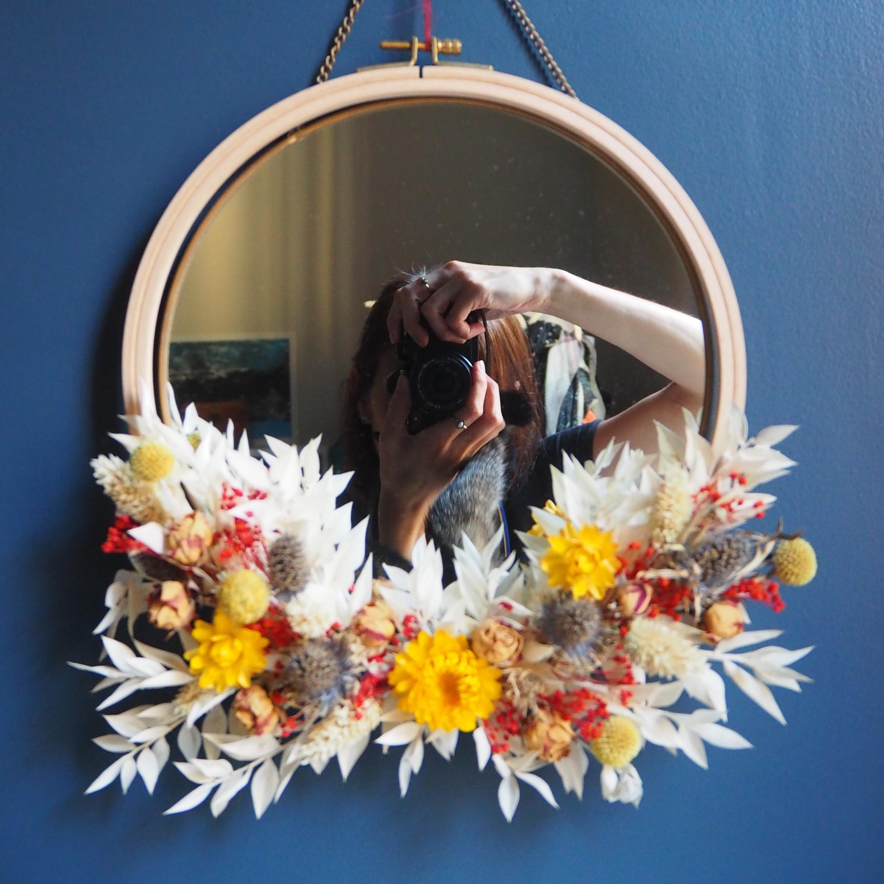 Auto-portrait d'une femme se prenant en photo devant un miroir rond, sur lequel est accroché une couronne de fleurs séchées faite à partir d'un tambour à broder en bois. Le miroir et la couronne sont accrochés sur un mur de couleur bleu. On peut également apercevoir dans le miroir la queue d'un chat tabby. Ambiance boho chic. Art botanique, cyanotypes originaux et tirages uniques. Art mural inspiré par la nature, pour une décoration d'intérieur boho chic, bohème, champêtre, végétal et eco-friendly.
