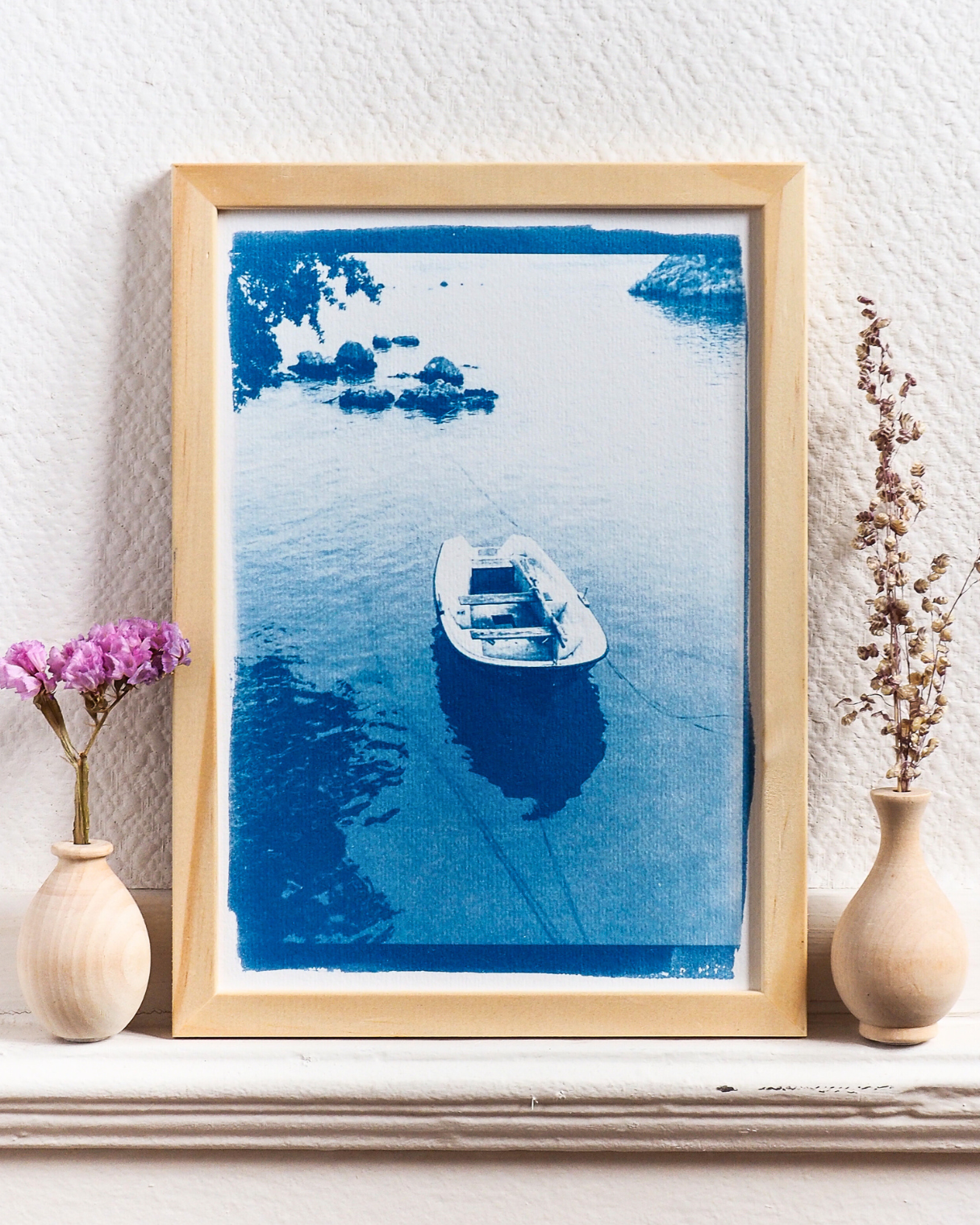 Photographie alternative développée avec la méthode de la cyanotypie, représentant une barque sur l'eau. Il y a des arbres dans le coin gauche en haut de l'image, qui se reflètent sur la surface de l'eau en bas à gauche. Le tirage est bleu de Prusse, ce qui donne une atmosphère onirique à la scène. Ce tirage est parfait pour une décoration boho chic et romantique, voire nostalgique et vintage, de votre intérieur.