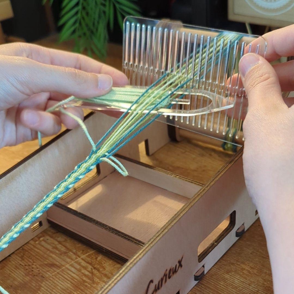 Photo d'un pendentif tissé aux couleurs trans et d'un porte-clés arc-en-ciel de la collection kaléidoscope queer posés dans l'herbe, à coté d'un packaging avec le logo de la marque