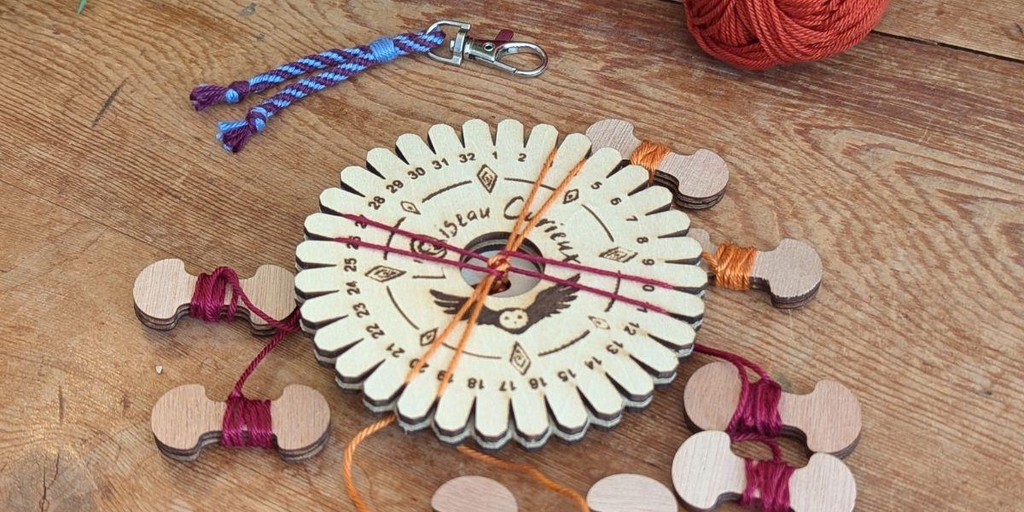 Disque de tressage posé sur une table, avec des fils orange et bordeaux en cours de tressage et un porte-clés tressé en arrière plan.