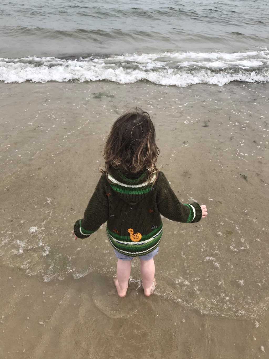 A child stood wearing a cardigan in the shallow water on a beach with their back to the photographer. 