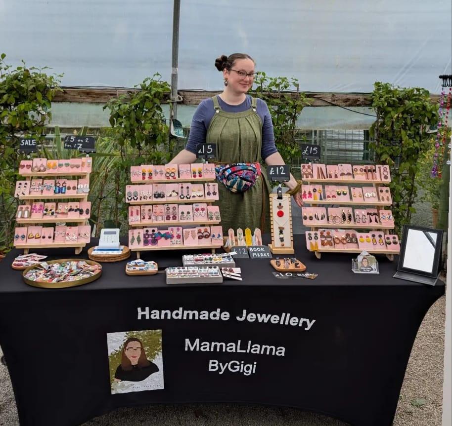 A woman named Gigi stands behind a table displaying a variety of handmade jewellery at a market stall. The table is covered with neatly arranged earrings, necklaces, and bookmarks on pink display boards. The stall is branded with a black tablecloth featuring the text "Handmade Jewellery MamaLlamaByGigi". Gigi is smiling and wearing a colourful fanny pack. The background includes greenery and a partially transparent cover, indicating an outdoor market setting.