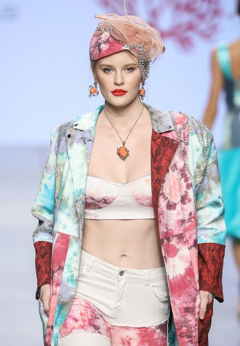 Modella in primo piano sulla passerella durante la Mercedes-Benz Fashion Week di Mosca, sfilata Coral Dream 2022 di Marita Balàzs, indossa cappotto oversize e pantaloni con stampa tie-dye nei toni corallo e turchese, top a fascia panna e turbante decorato con applicazioni gioiello.