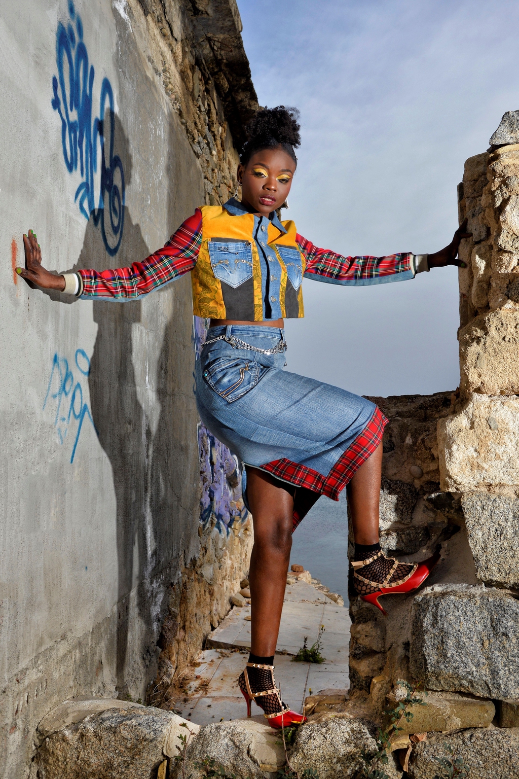 Modella in posa tra le rovine di un edificio in pietra, indossa un look Marita Balàzs composto da giacca corta patchwork in denim, tartan rosso e giallo, camicia a quadri e gonna midi in denim con inserti tartan. Ai piedi, sandali neri decorati e calzini a rete. Sullo sfondo, muro con graffiti e cielo nuvoloso.