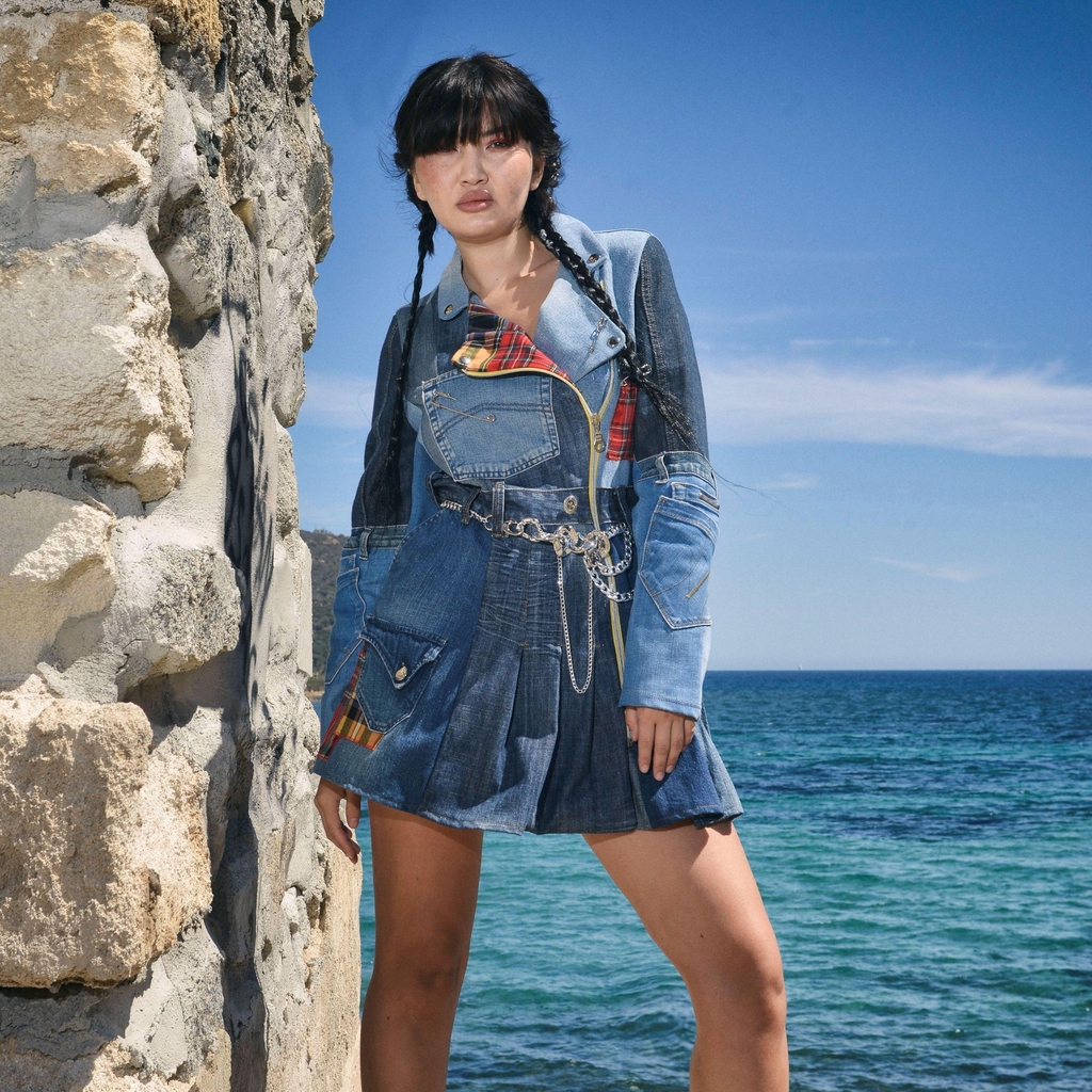 Modella in posa accanto a un muro di pietra, con il mare turchese sullo sfondo, indossa un abito corto Marita Balàzs in denim patchwork con dettagli tartan, cintura a catena e stivaletti neri con plateau. Lo styling è grintoso e contemporaneo.