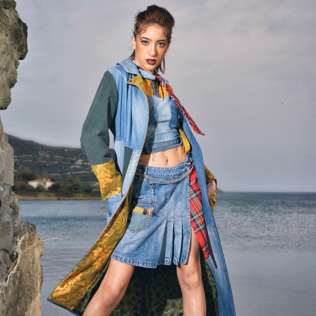 Modella in posa su una scogliera davanti al mare, indossa un trench Marita Balàzs in denim patchwork con dettagli verdi e fodera animalier, top corto in denim, minigonna a pieghe con inserti tartan e cintura rossa, calzini e sandali verdi. Sullo sfondo, paesaggio costiero e cielo nuvoloso.