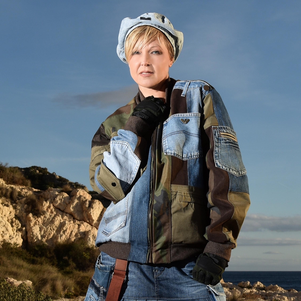 La stilista Marita Balàzs in posa all’aperto davanti a una scogliera, indossa una giacca patchwork in denim e tessuto mimetico, una gonna di jeans plissettata, guanti neri e cappello coordinato. Sullo sfondo si vede il mare e la vegetazione costiera.