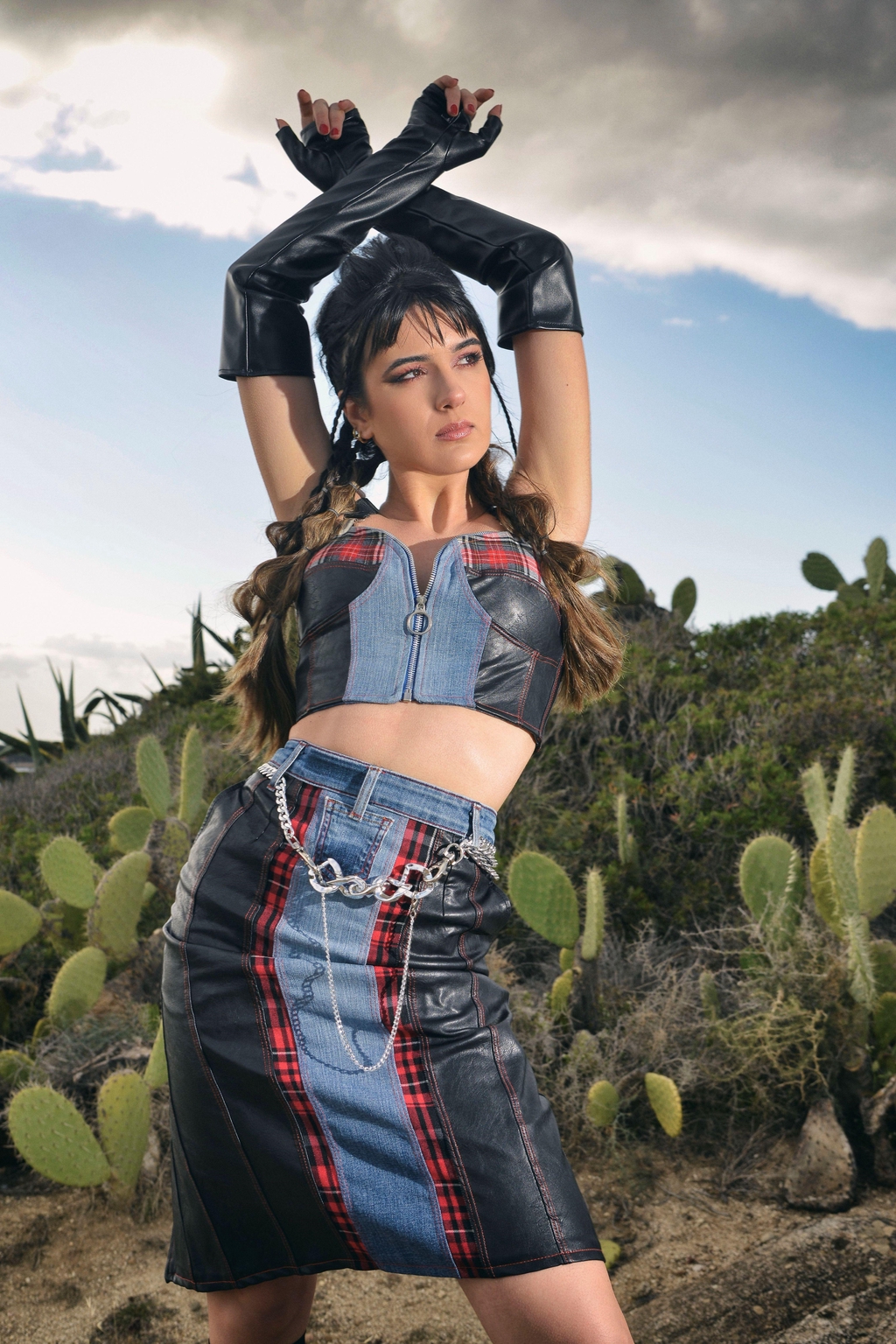 Modella in posa tra i cactus sotto il cielo nuvoloso, indossa un completo Marita Balàzs composto da top corto e gonna a tubino in patchwork di pelle nera, denim e tartan rosso, con cintura a catena e guanti lunghi in pelle nera. Lo styling è grintoso e dallo spirito punk.