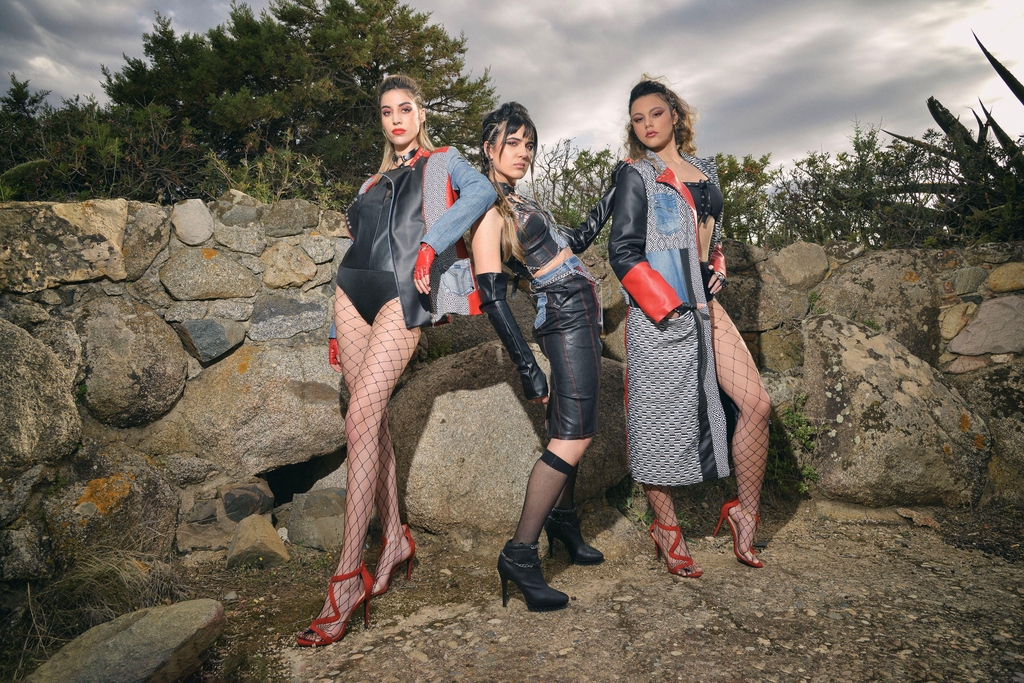 Tre modelle posano all’aperto davanti a un muro di pietra e vegetazione, indossando look Marita Balàzs in stile punk-chic: body neri con giacche in denim e pelle rossa, pantaloni in ecopelle, trench patchwork, calze a rete e scarpe rosse con tacco. L’atmosfera è grintosa e decisa.