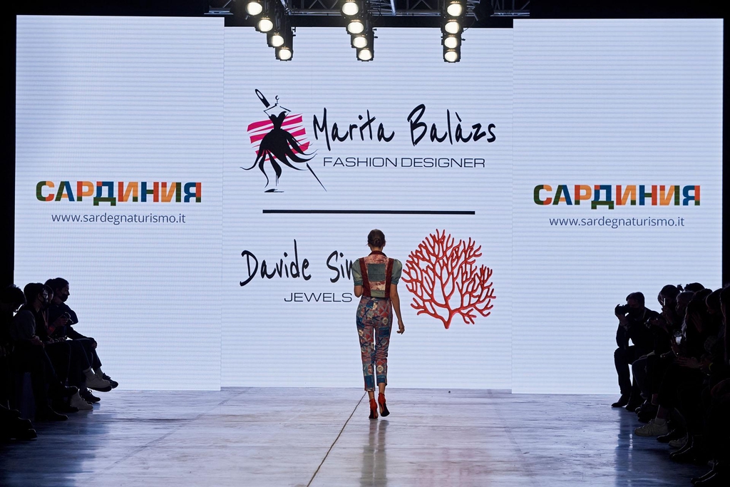 Modella di spalle sfila su una passerella illuminata, indossa una tuta lunga aderente con stampa colorata e dettagli cut-out sulla schiena. Sullo sfondo, maxi schermo con logo “Marita Balàzs Fashion Designer” e pubblico seduto ai lati della passerella.