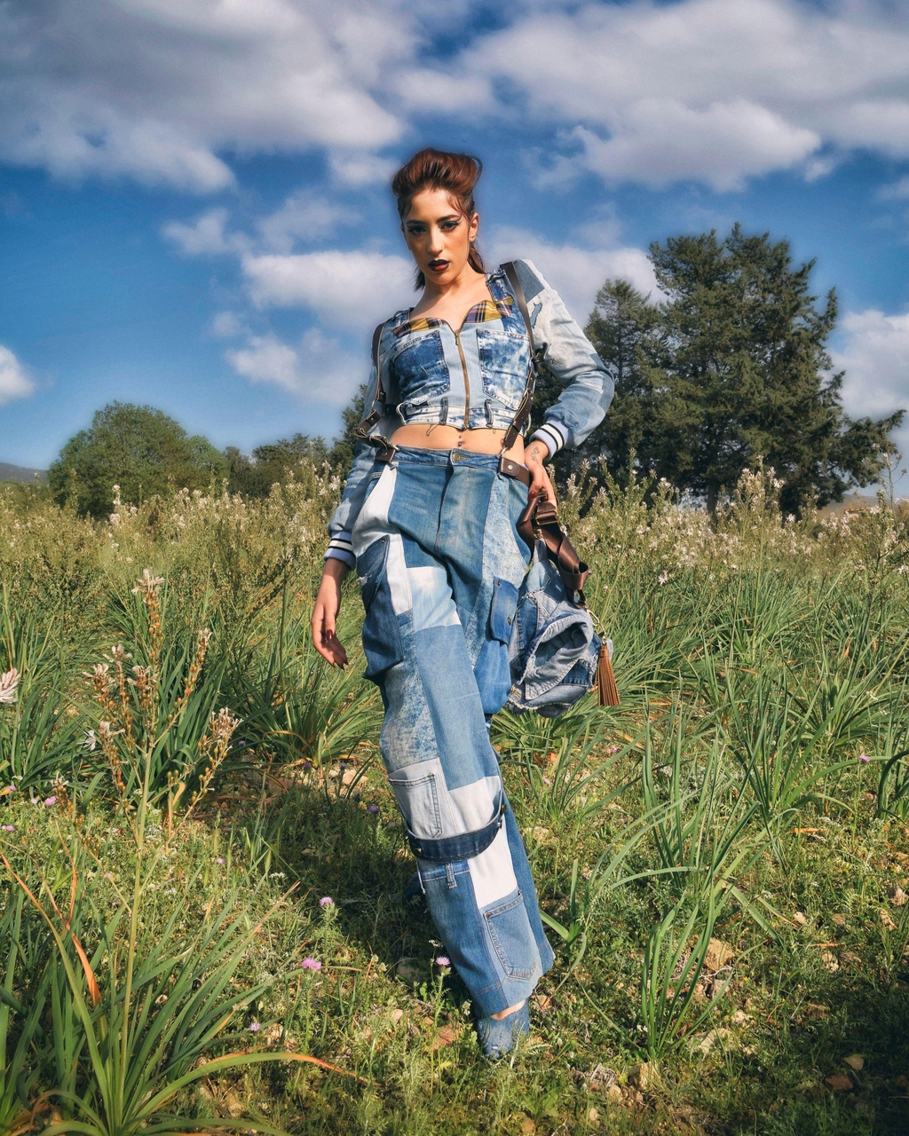 Modella in piedi in un prato verde sotto un cielo azzurro con nuvole, indossa un completo Marita Balàzs in denim patchwork composto da giacca corta e pantaloni a vita alta, crop top coordinato e borsa in denim. Lo styling è grintoso e contemporaneo.
