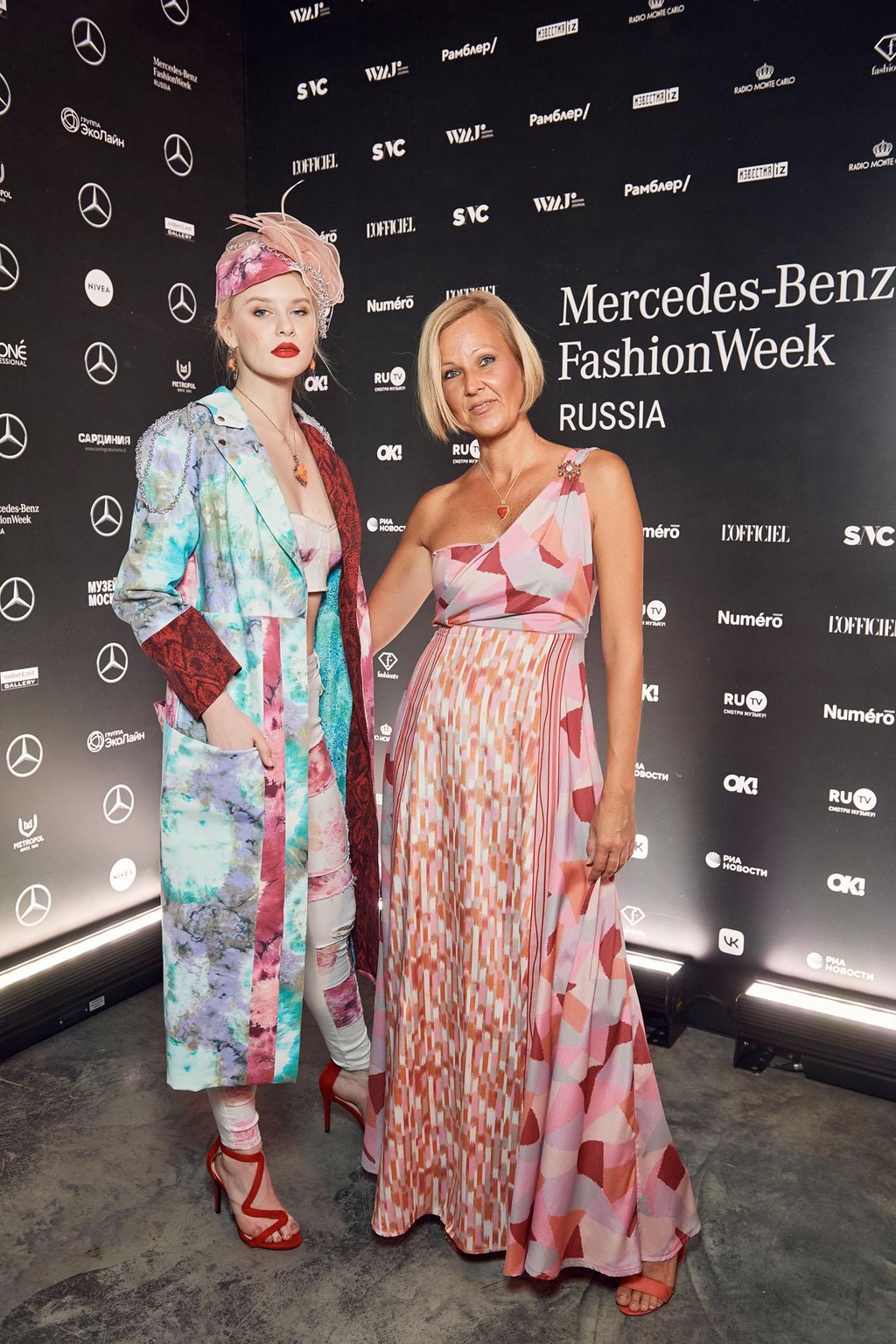 La stilista Marita Balàzs e la sua modella posano davanti a un pannello nero con loghi della Mercedes-Benz Fashion Week Russia. Una indossa un lungo cappotto colorato con motivi geometrici e sandali rossi, l’altra un abito lungo monospalla rosa con motivi astratti.