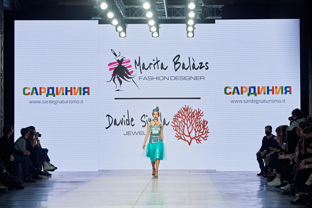 Modella sfila su una passerella illuminata, indossa una gonna ampia in tulle verde acqua, top bianco con dettagli argentati e una giacca corta stampata, sandali argento e cerchietto decorato. Sullo sfondo, maxi schermo con logo “Marita Balàzs Fashion Designer” e pubblico seduto ai lati della passerella.