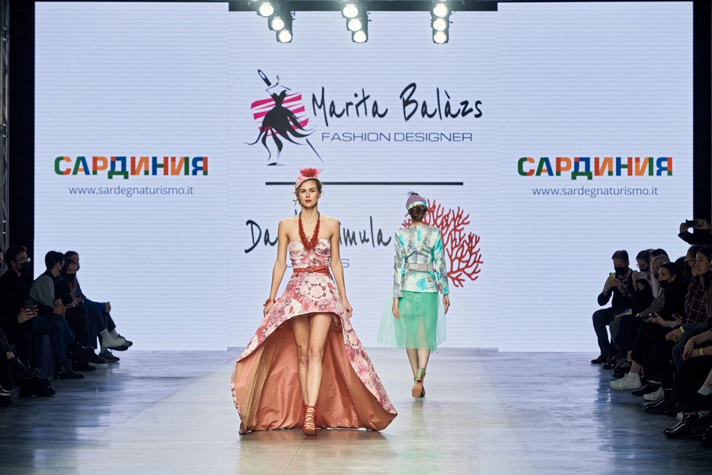 Modella sfila su una passerella indossando un abito lungo rosa con stampa floreale, corpetto aderente e ampia gonna con spacco frontale, cappellino decorato e sandali rosa; sullo sfondo, un’altra modella con abito verde acqua e giacca stampata. Dietro, maxi schermo con logo “Marita Balàzs Fashion Designer” e pubblico seduto ai lati della passerella.