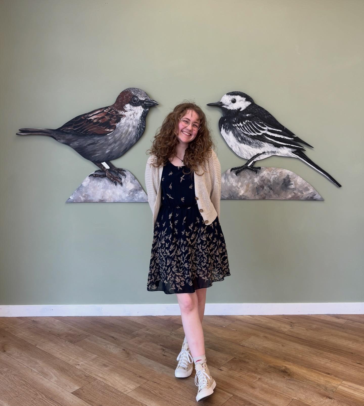 Artist Amy Foster - Young woman with curly hair wearing a dress with birds on standing smiling in front of two large bird artworks hanging on a green gallery wall