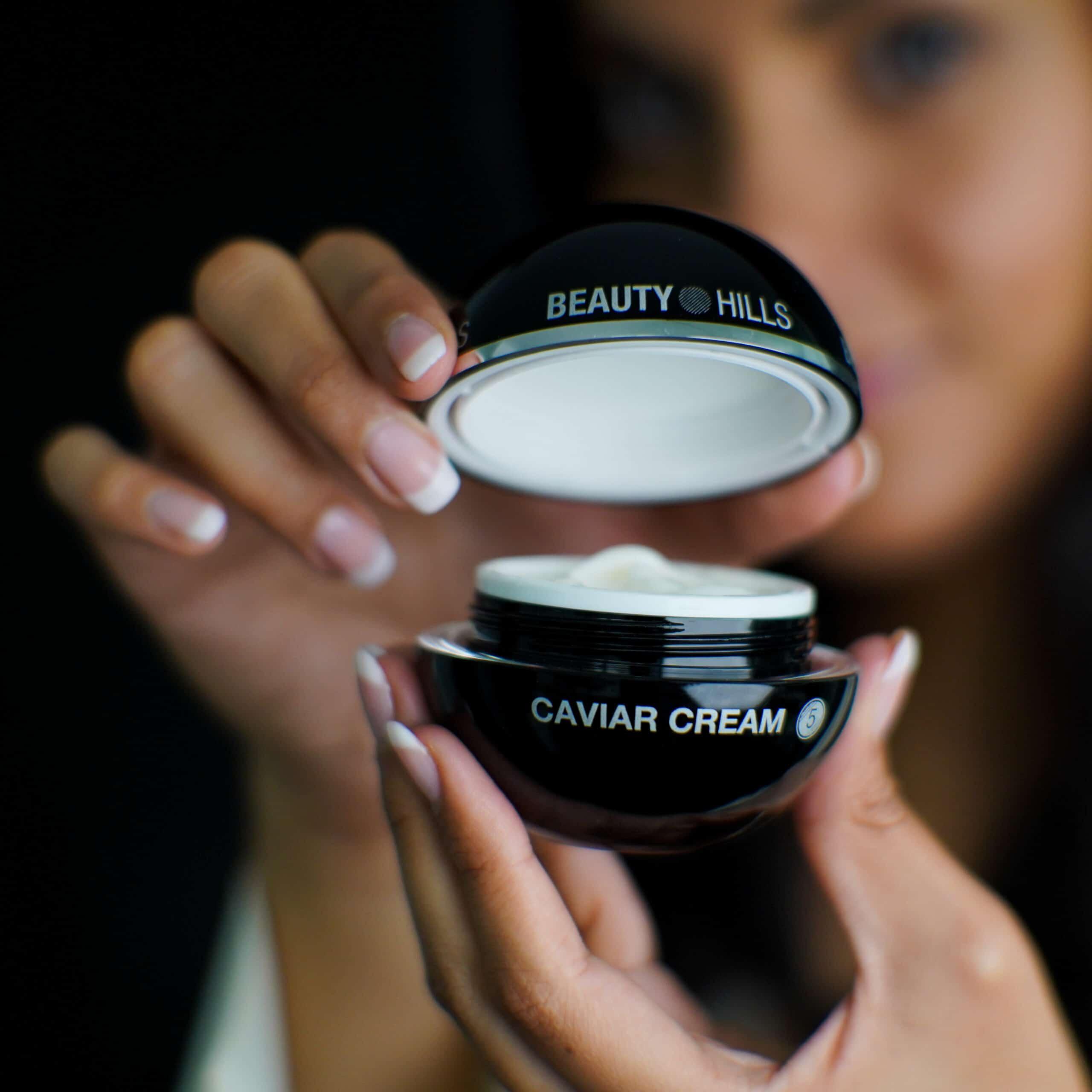 Auf dem Bild ist eine geöffnete, schwarze, kugelförmige Caviar Creme Schachtel in den Händen einer Frau zu sehen. Der Markenname 'Beauty Hills' ist gut sichtbar auf der Schachtel aufgedruckt. Das Bild präsentiert die elegant verpackten System-Kosmetikprodukte von Beauty Hills, die im Shop angeboten werden.