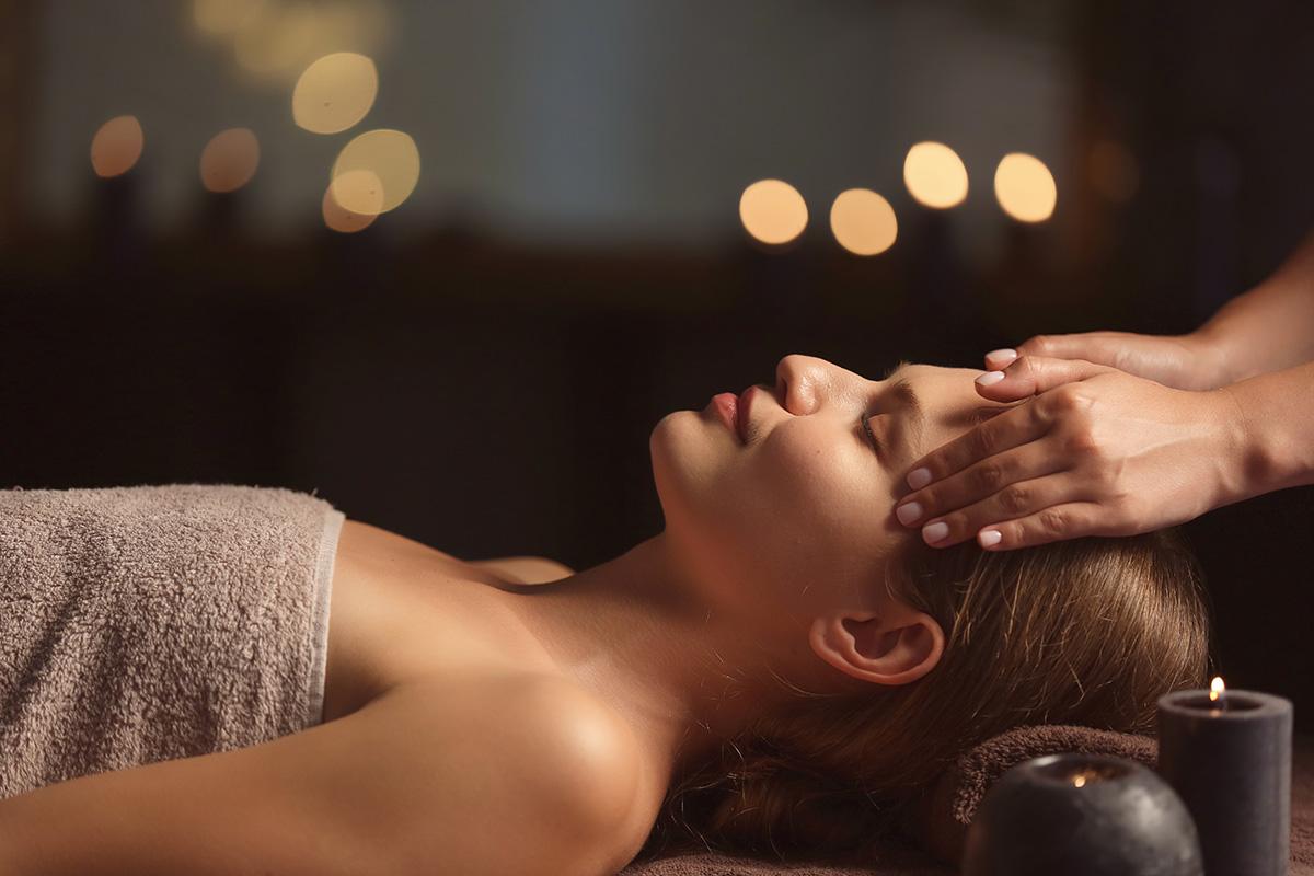 Eine junge Frau liegt entspannt auf einer Kosmetikliege und erhält eine wohltuende Gesichtsmassage. Ihre halb geschlossenen Augen und das leichte Lächeln auf ihrem Gesicht spiegeln tiefe Entspannung und Wohlbefinden wider. Die Hände der Masseurin sind bei der Gesichtsmassage zu sehen. Im Hintergrund sorgen brennende Aroma-Kerzen für eine ruhige, angenehme Atmosphäre, während die Massage die Haut, das Bindegewebe im Gesicht, Hals und Dekolleté gesund und straff pflegt.
Dieser Text hebt die entspannende Wirkung und die positiven Effekte auf die Haut hervor.