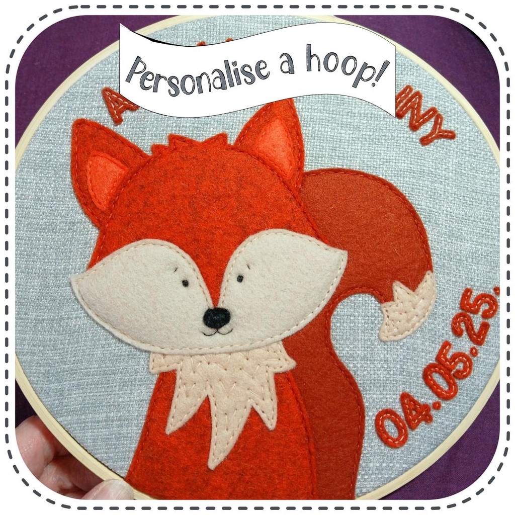 A fox sewn on in wool felt. He is in an embroidery hoop with personalised text added which is also sewn on in wool felt. The digital text reads 'Personalise a hoop!' The fox is smiling and has a cheeky looking face. His colours are a rich orange and warm paprika brown with beige detail on his tummy, face and tail.