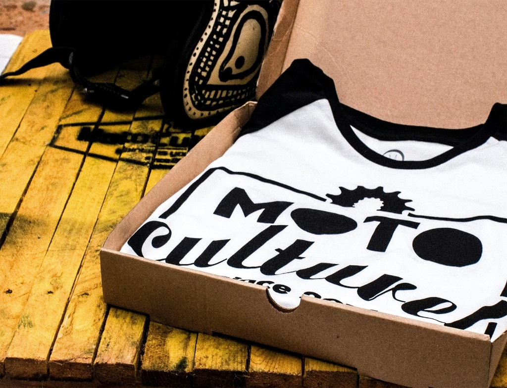 T-shirt blanc avec logo Moto Culture plié dans une boîte en carton, prêt pour expédition, sur une table en bois jaune avec sac à dos noir à côté