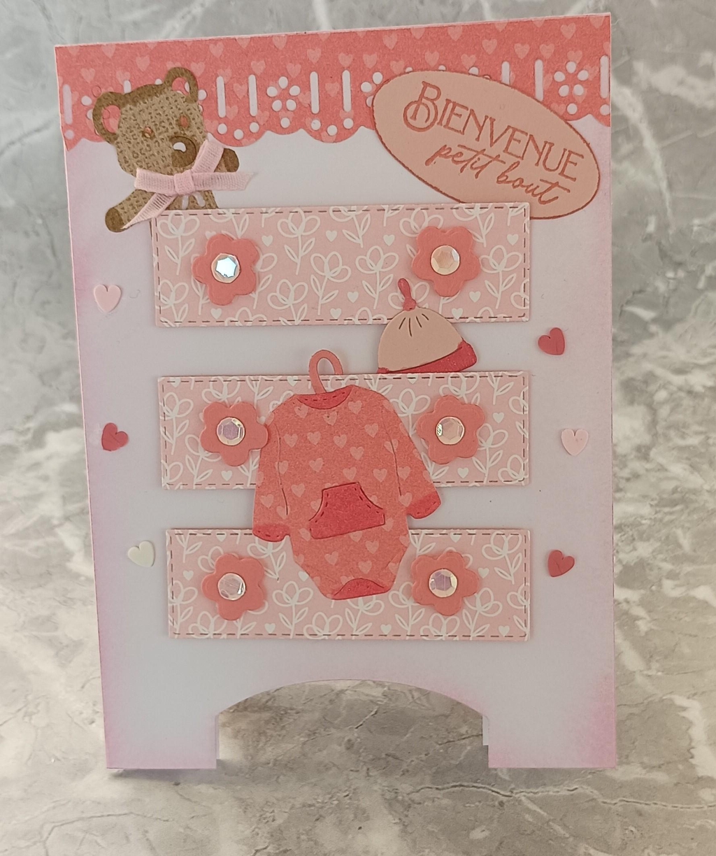 Carte de naissance fille faite main en scrapbooking rose pastel, décorée d'un ourson, d'un body de bébé en relief et de l'inscription 'Bienvenue petit bout'."