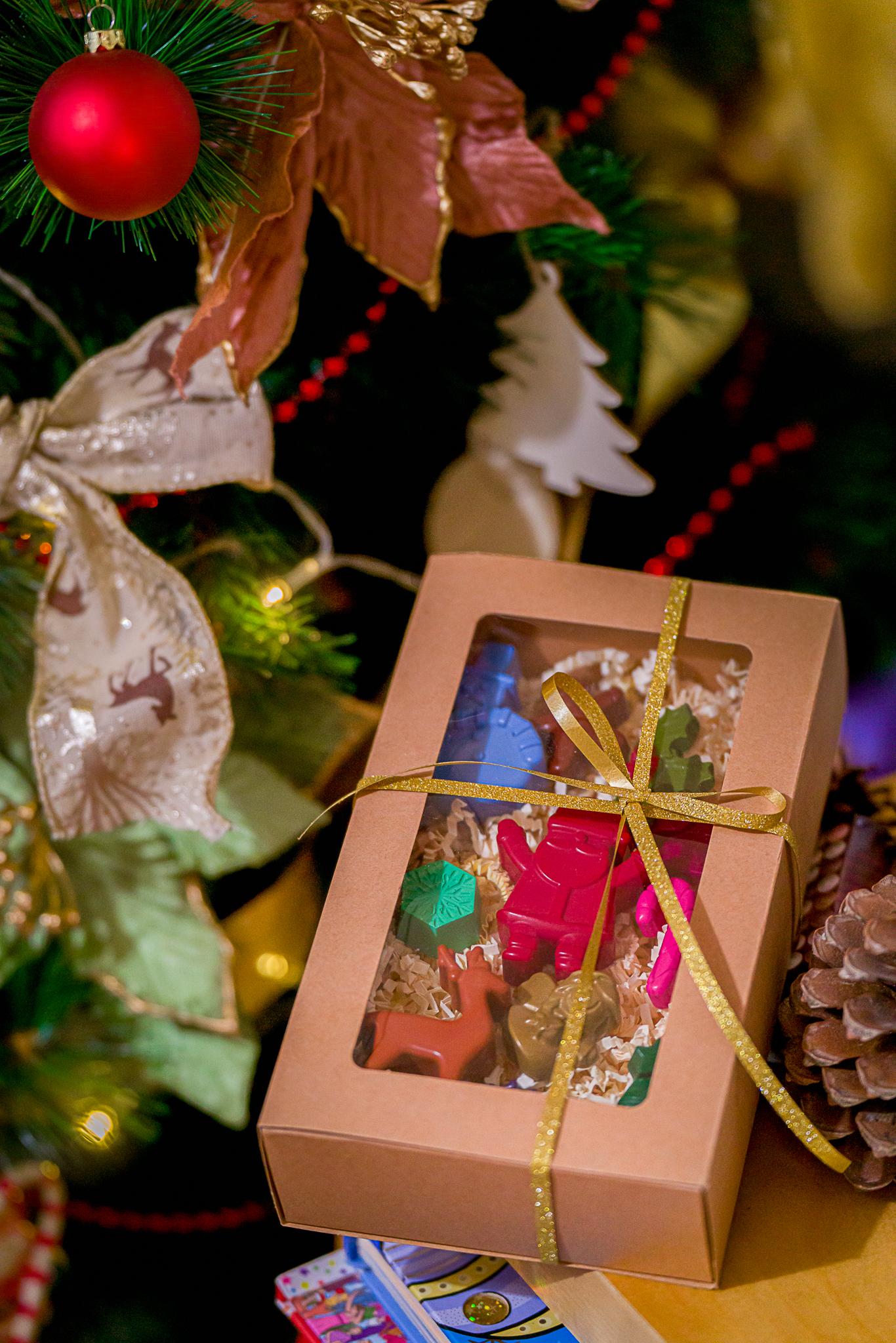 boîte carton kraft à fenêtre placée sous le sapin de Noël et contenant de crayons en cire aux figures de Noël et décoré d'un ruban doré
