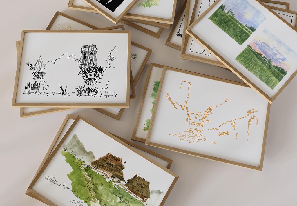 Stos printów przedstawiających odręczne szkice z podróży, w różnych technikach takich jak akwarela, kredka i piórko, oprawione w drewniane ramki. Obrazki ułożone jeden na drugim, ukazujące różnorodne sceny i budynki.