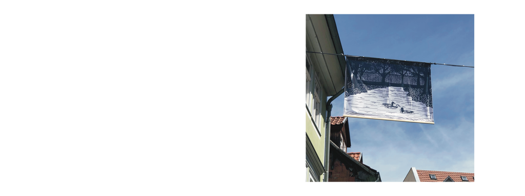 Große schwarz-weiße Fahne mit einem Kunstwerk, das zwei Enten auf einem See zeigt, aufgehängt an einem Seil zwischen zwei Gebäuden.