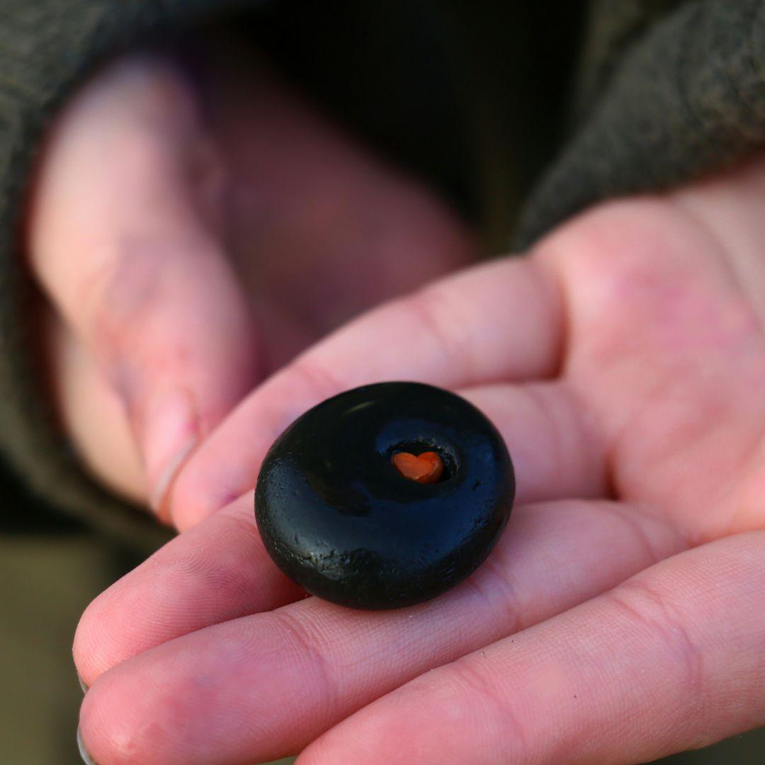 Photo de deux mains jointes tenant un caillou noir avec un petit cœur rouge au centre, symbolisant l’engagement de la créatrice à reverser une partie de ses ventes à une association de protection de la nature et de l'environnement. Cette image représente l’acte de solidarité et le lien intime entre chaque création de bijoux et la mission de préserver notre planète