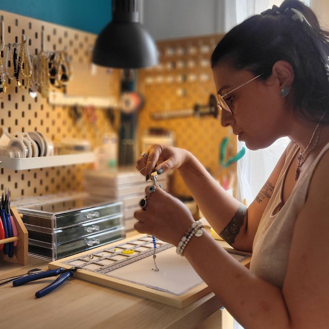 Photo de profil de la créatrice de bijoux artisanaux en plein travail, concentrée sur l’assemblage minutieux de bracelets faits main. Elle manipule des outils de bijouterie dans son atelier, où chaque pièce prend vie à partir de pierres naturelles, de perles et de métaux soigneusement sélectionnés. Une plongée dans l’univers d’une marque française inspirée par la nature et portée par un savoir-faire authentique