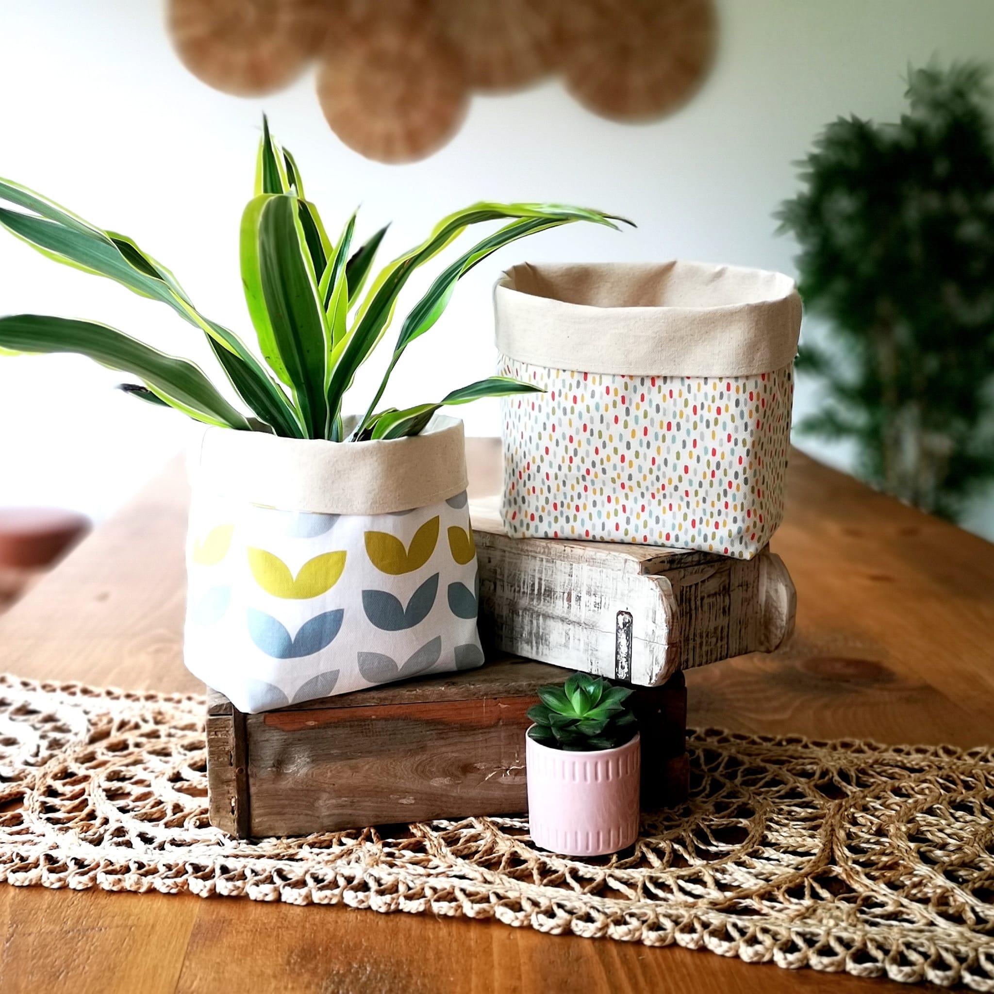 Two Sew She Did fabric baskets made using designer textile remnants atop a wooden table. One is white with a green. blue and grey leaf design and houses a leafy house plant. The second is made from natural cotton fabric with multi-coloured dashes. There is a small succulent plant in the foreground in a pink pot.
