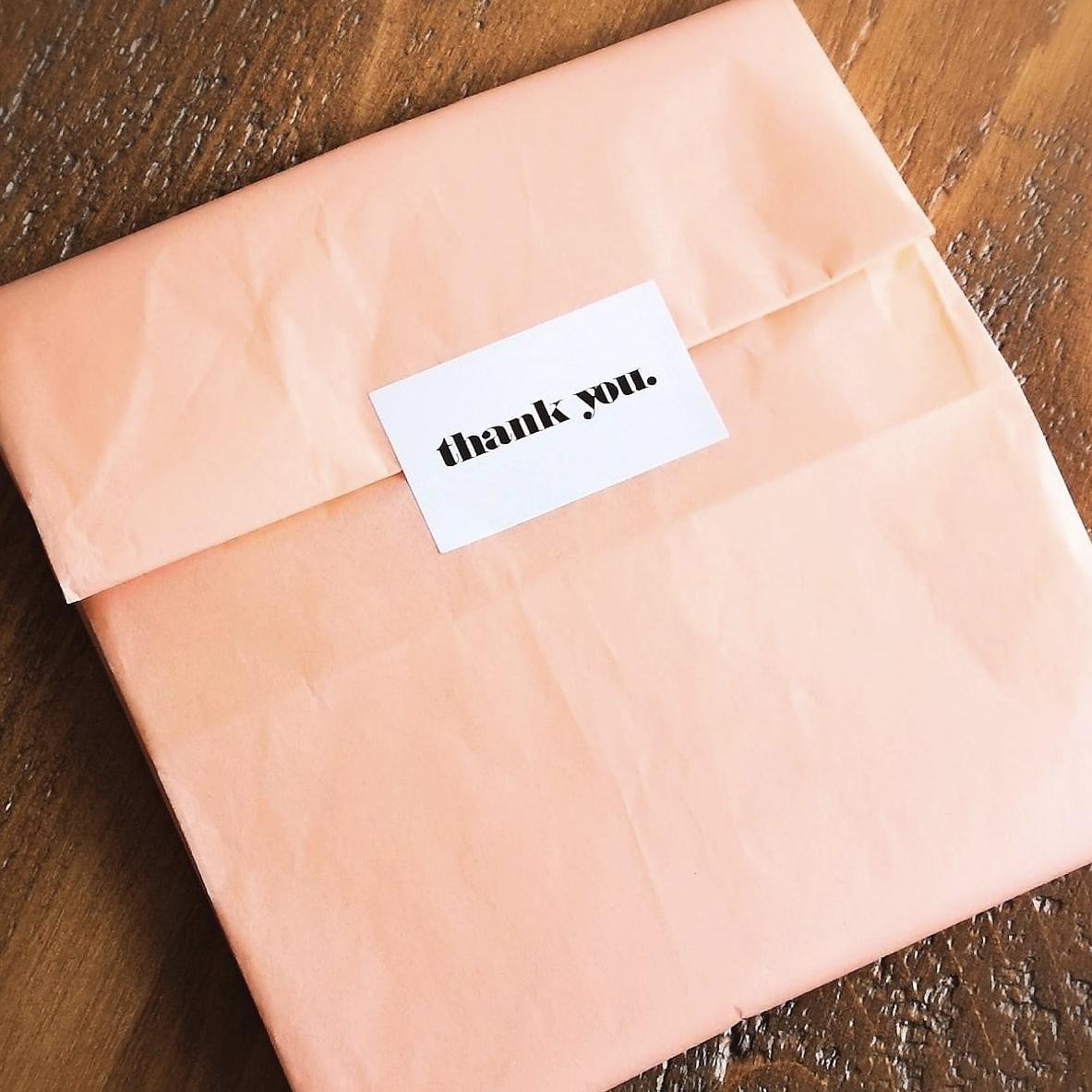 A table with a gift on it wrapped in peach tissue paper. There is a sticker with "thank you" sealing it closed.