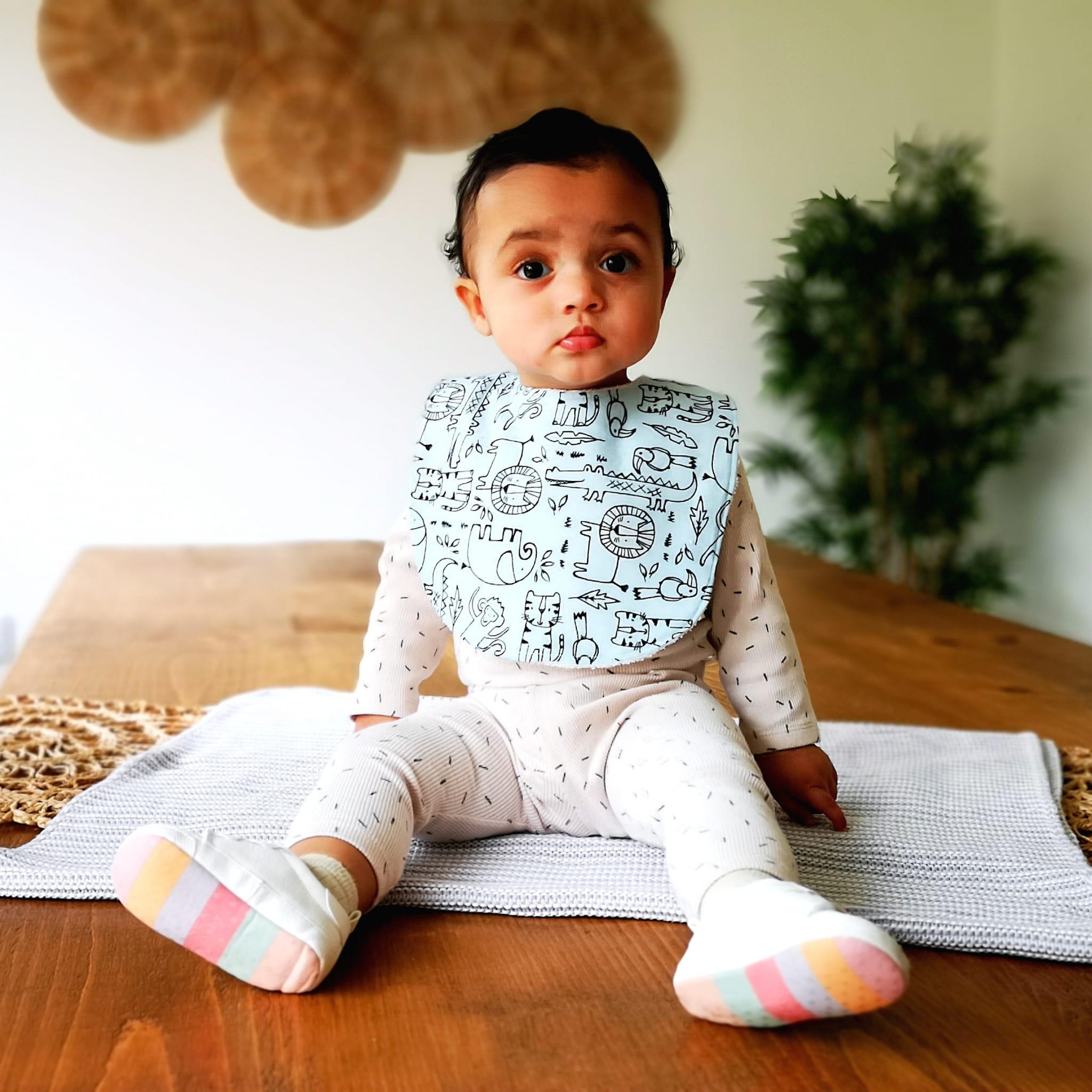 A baby sat on a blanket on a wooden table wearing a baby bib. The bib is blue with wild animals on it. There is a plant in the background.